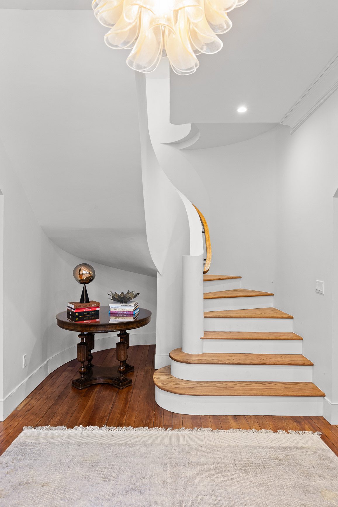 Curved plaster staircase with white oak treads rising under a sculptural chandelier